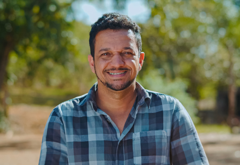 A man wearing a blue checkered button-down collared shirt smiles at the camera while standing in an open field with trees behind him.