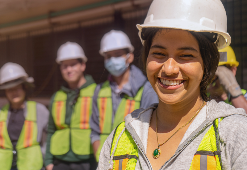 Uma jovem trabalhadora usando um capacete de proteção sorri, com vários outros trabalhadores em pé atrás dela.