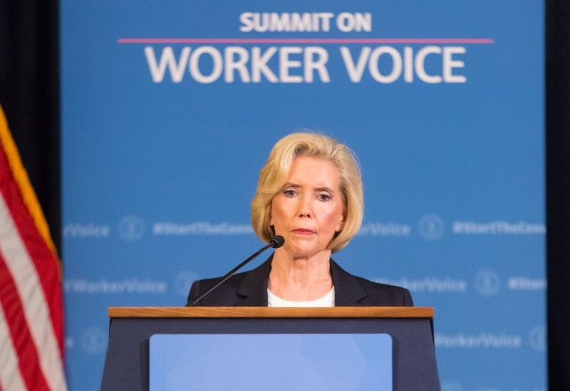 Lilly Ledbetter, an older white woman with short blond hair, is pictured from the shoulders up behind a podium. A blue screen behind her bears the words “Summit on Worker Voice.”