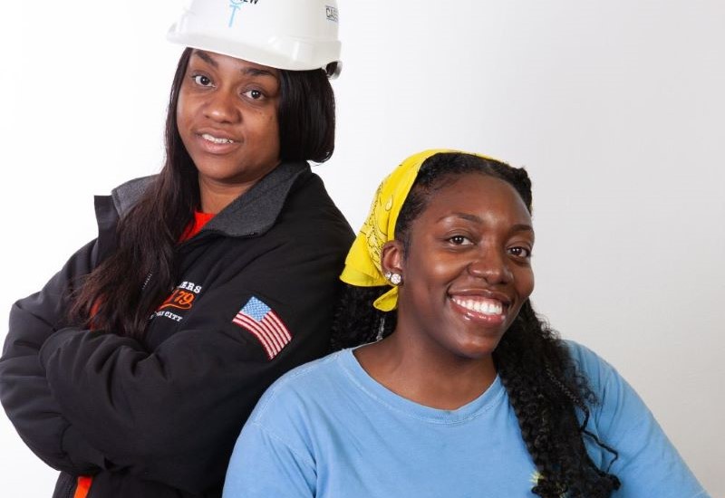 A Black woman with long hair standing while wearing a black jacket and white hardhat and a Black woman with braids wearing a yellow scarf and blue t-shirt resting her arm on a white hardhat.