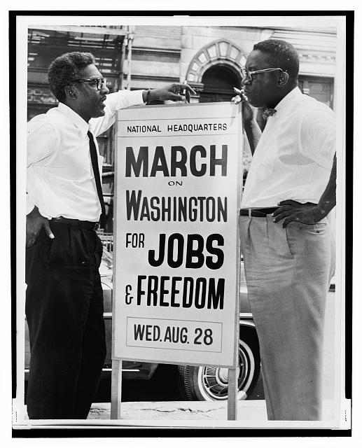 Black and white photo from the Library of Congress shows Bayard Rustin and Cleveland Robinson, two Black, male March organizers, speaking by a sign reading “March on Washington for Jobs and Freedom, Wed. Aug. 28”