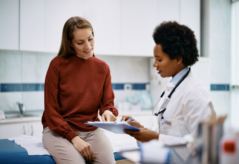 A woman wearing a rust-colored turtleneck and khaki pants reviews medical documents on a clipboard with a doctor wearing a white coat and stethoscope. 