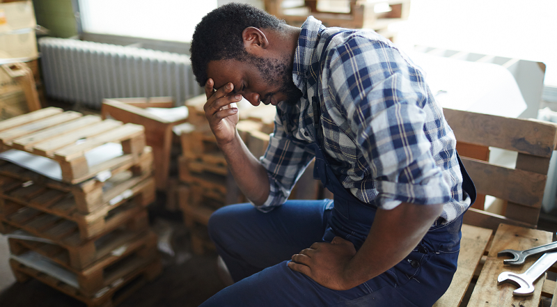 A middle-aged worker in a checked shirt sits in a room full of pallets and tools, holding his head in his hand with a stressed expression on his face.