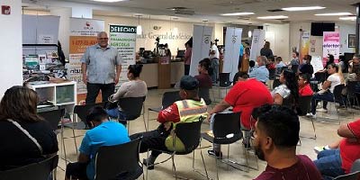 A man delivers a presentation to a group of people at the Mexican Consulate. A banner with information about EMPLEO is behind him.