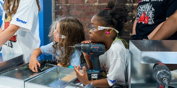 A young camper listens to an adult speak, her chin resting on a power drill, at Tools & Tiaras.