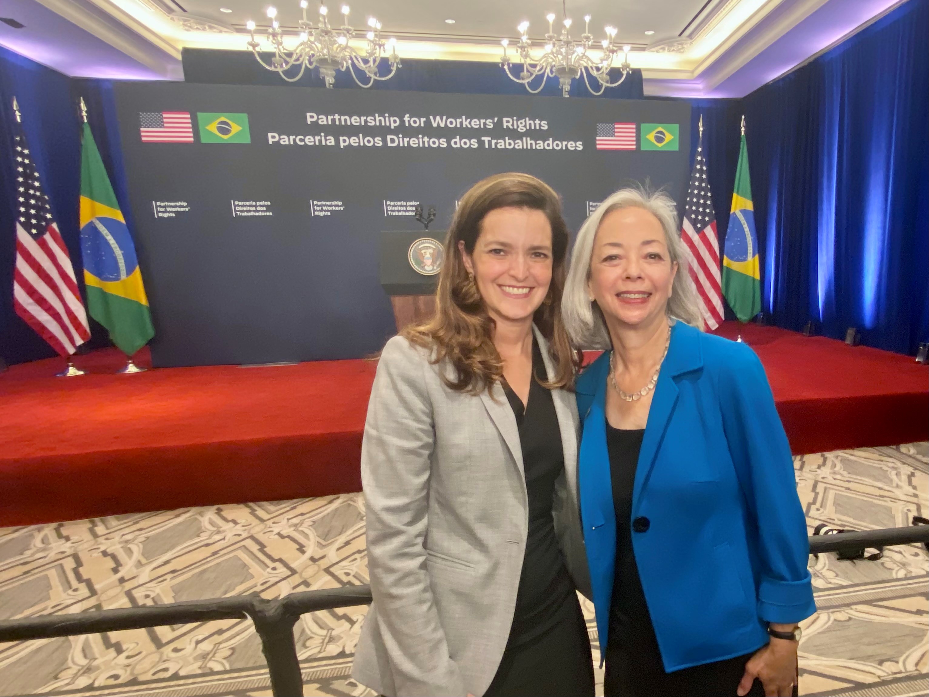 Thea Lee and Special Representative for International Labor Affairs Kelly Fay Rodriguez stand in front of the stage that reads, "Partnership for Workers' Rights," flanked by the U.S. and Brazilian flag.
