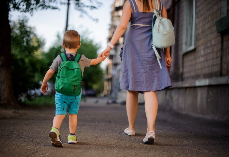 A rear view of a mom wearing a blue dress and flat shoes walking with her son wearing a grey shirt, blue shorts, sneakers and a green backpack while holding hands.