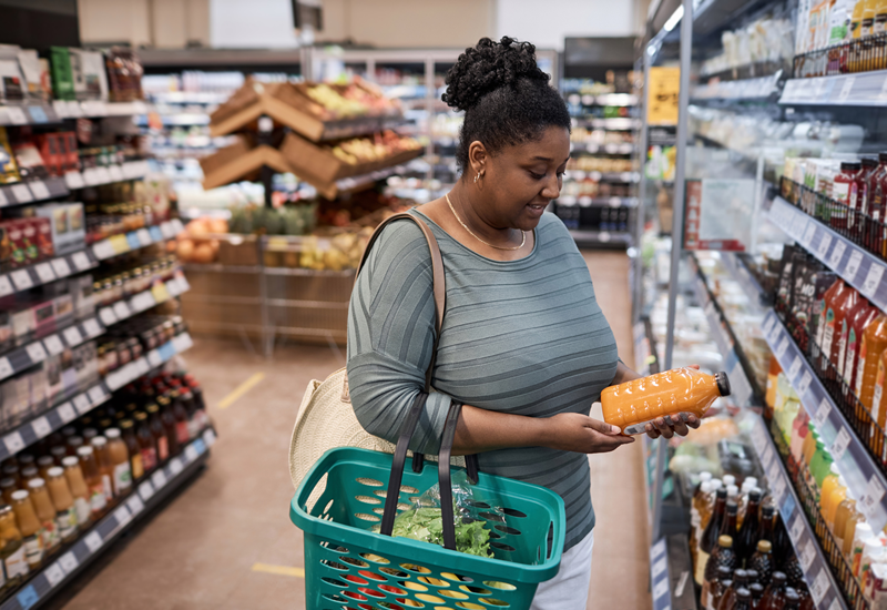 一名妇女提着购物篮，在杂货店的冰箱过道上看着一瓶果汁。