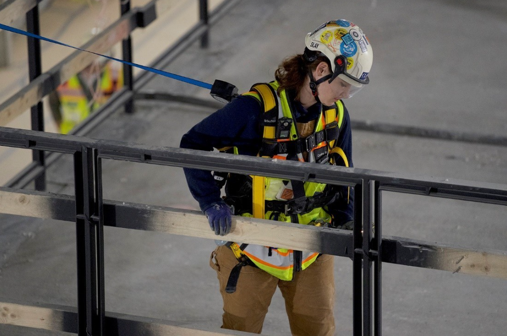 Uma trabalhadora da construção civil vestindo equipamento de alta visibilidade, capacete de segurança com faixa de queixo e viseira transparente, arnês de proteção contra quedas e luvas.