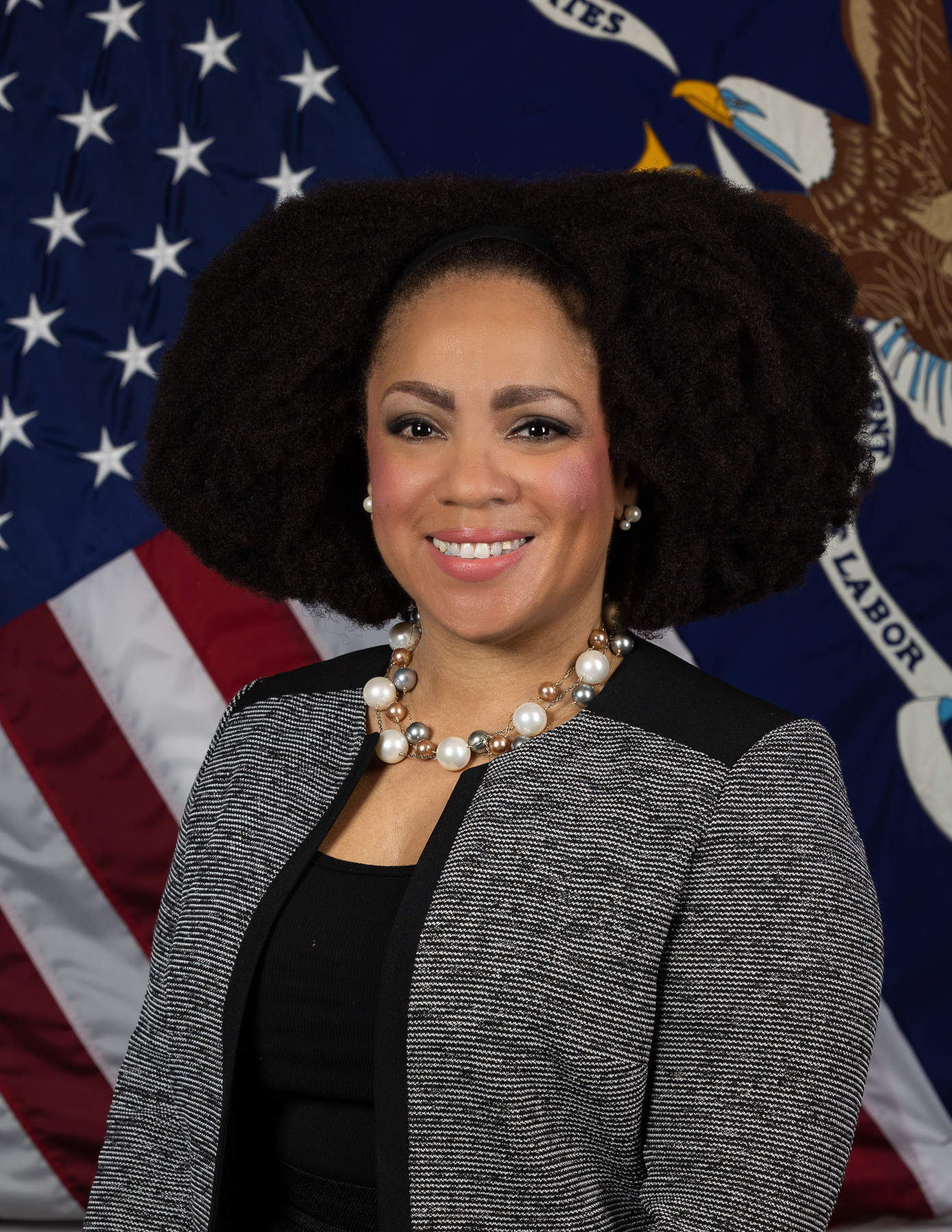 En la imagen, una mujer afroamericana con el pelo por los hombros sonríe a la cámara delante de la bandera del Departamento de Trabajo y de la bandera de los Estados Unidos. 