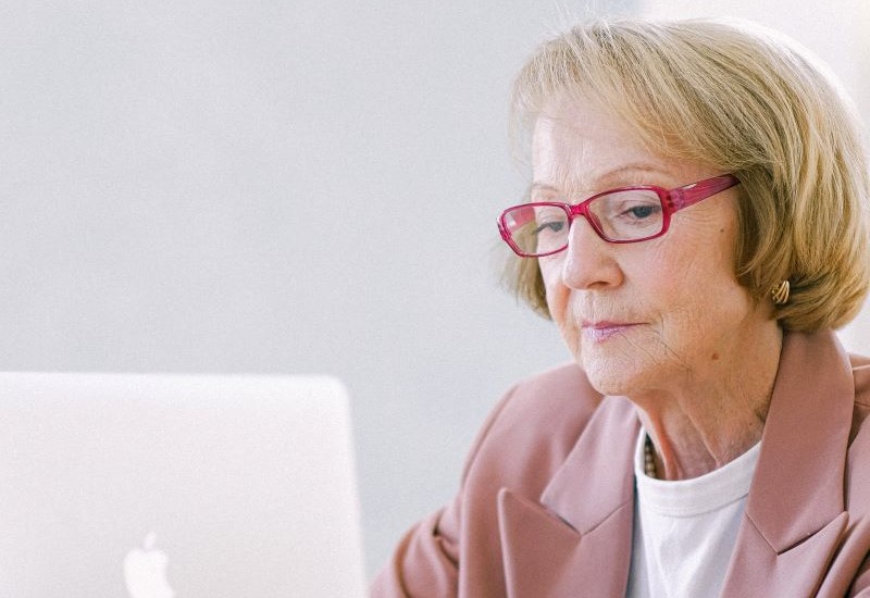 An older white woman with short blond hair is shown from the shoulders up. She wears red glasses and is looking at a laptop.