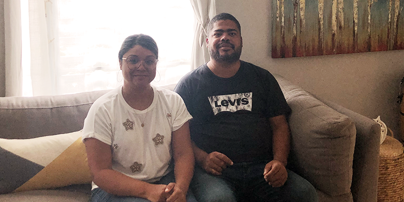 A man and woman sit on a couch in a living room, smiling at the camera.