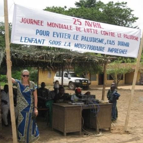 A young American woman in sunglasses stands by a group of people working behind desks in a dirt clearing. A sign in French advertises World Day Against Malaria.