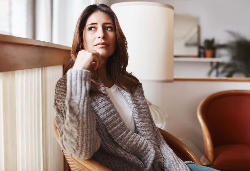 A woman sits in a living room with her head resting on her fist, looking pensively into the distance.