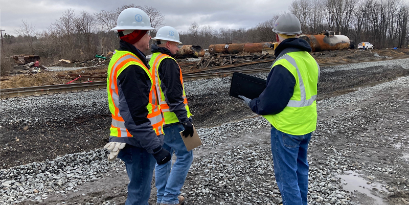 Three OSHA employees wearing hardhats and visibility vests survey the scene of the derailment.