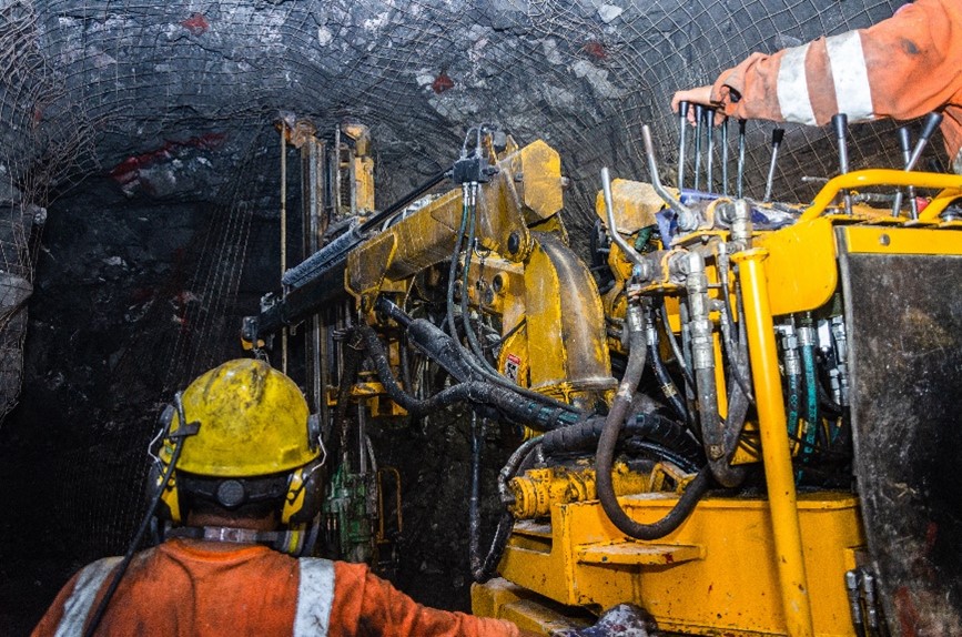 Miners are working in an underground mine under a roof supported with a mesh screen. 