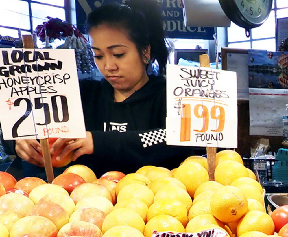 A young woman working at a fruit stand, advertising prices for apples and oranges.
