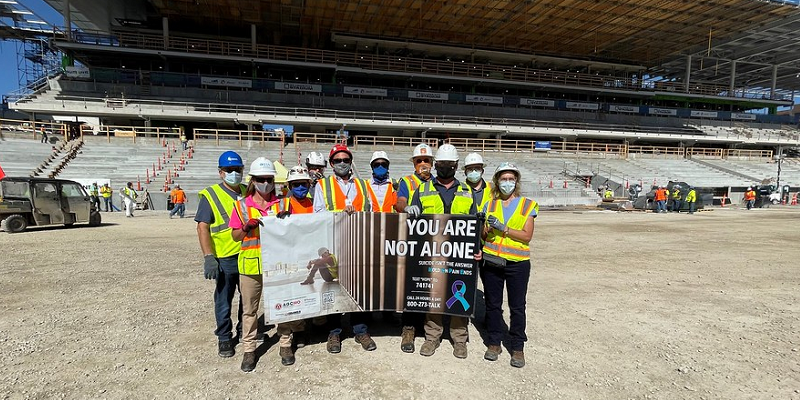 A group of construction workers standing in a stadium hold a banner that says "You are not alone."