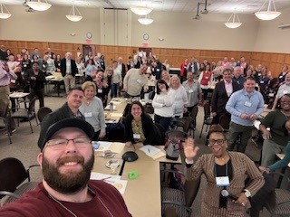 Un grupo de personas reunidas alrededor de mesas en una gran sala sonríen para una foto tipo selfie.