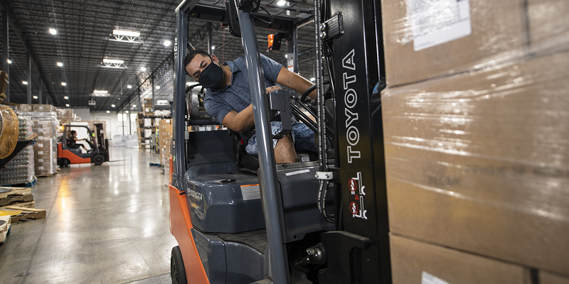 A male worker wearing a mask drives a forklift in a large warehouse