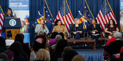 Wendy Chun-Hoon speaks behind a podium. A panel of speakers is seated to her left, in front of a full auditorium.