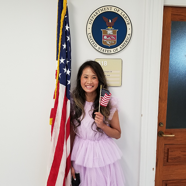 A woman in a pink dress poses by an American flag, holding a miniature flag and beaming at the camera. The Labor Department seal is visible behind her.