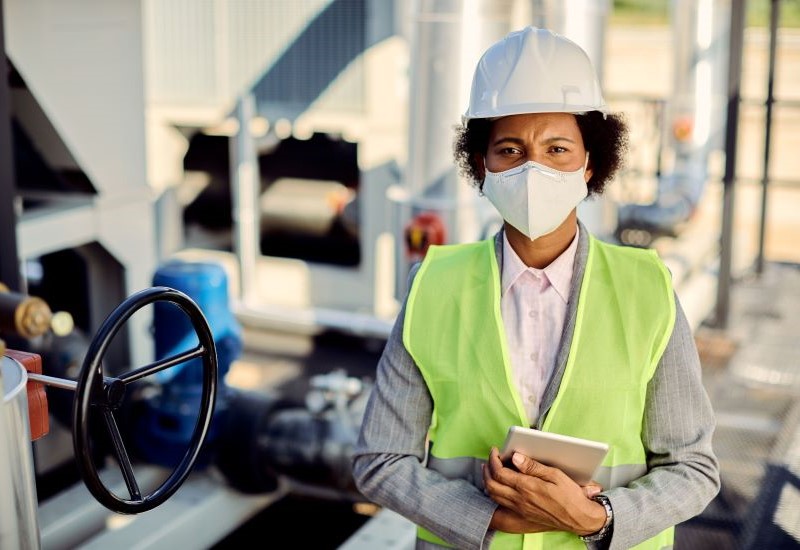 A middle-aged Black woman in a suit, safety vest, mask and hardhat poses at an industrial site.