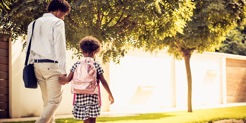 A dad carrying a briefcase walks his young daughter to school