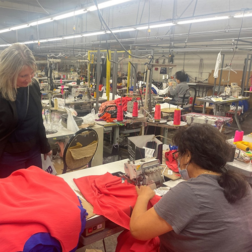 In a crowded workroom, a woman pushes red fabric through a sewing machine.