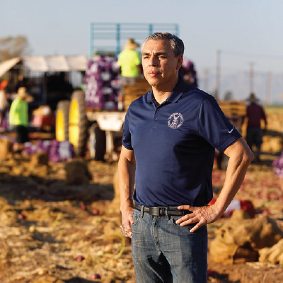 Wage and Hour investigator Juan Coria stands in a field. Workers are visible on a tractor behind him.