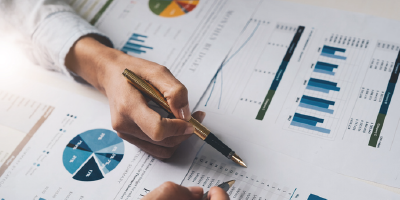 Close-up of hands holding pens above printouts of pie charts, bar charts and other data graphs. 