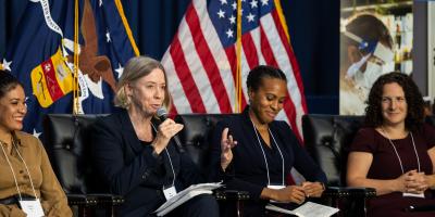 A seated woman in a business jacket addresses the audience. Three other women on the panel listen attentively.