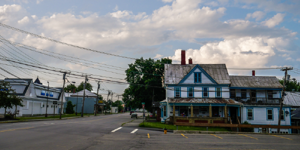 Una tranquila escena callejera en un pequeño pueblo rural que muestra una casa antigua de dos plantas con pintura azul y blanca desgastada bajo un cielo parcialmente nublado, con cables de electricidad que se entrecruzan por encima.