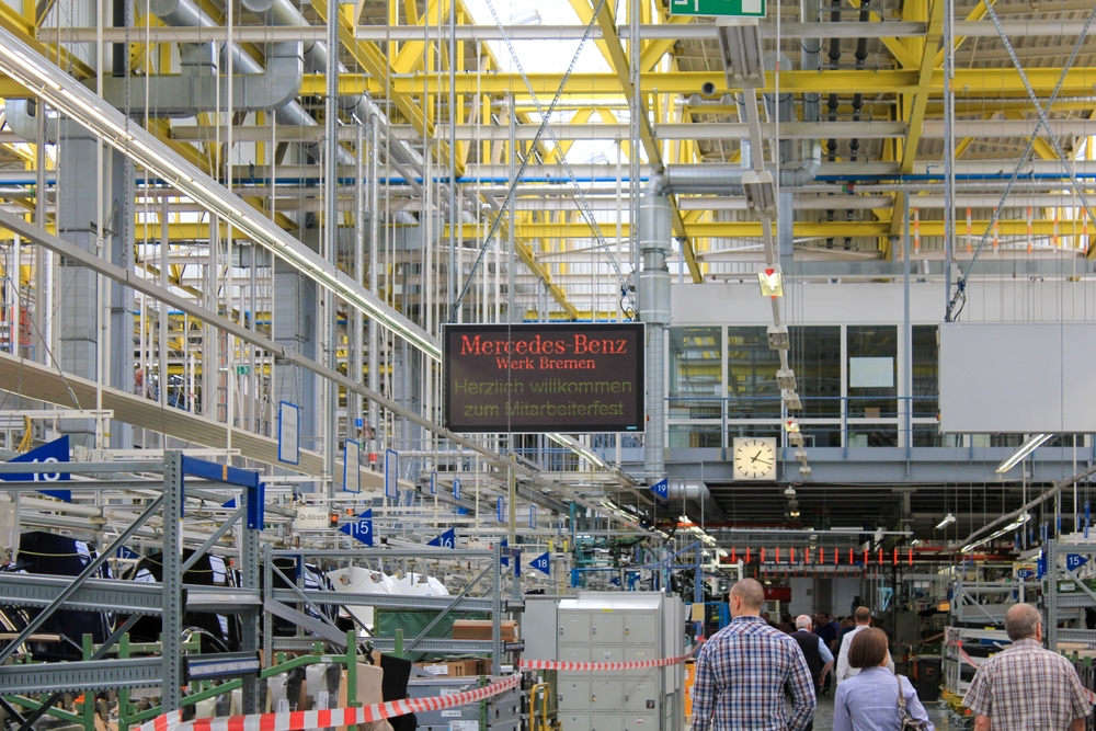 Several people in plain clothes walk through an auto factory. A sign in Germany reads “Mercedes-Benz Bremen plant, welcome to the employee party.”