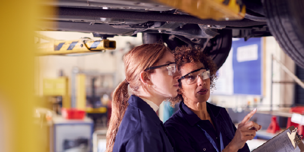 Two women in mechanics' coveralls examine a car on a lift.