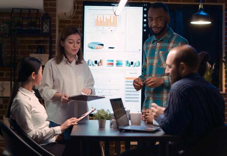 Several employees work around a table with laptops and tablets. A large screen with a data presentation is behind them.