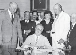 Black and white photo: President Franklin Roosevelt signs the Social Security Act, flanked by supporters, including Secretary Frances Perkins.