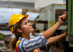 A woman wearing a hard hat works in a machine shop or industrial environment