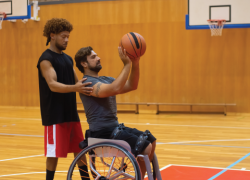 A personal trainer guides a man in a wheelchair as he prepares to shoot a basketball. Photo by Kampus Production.