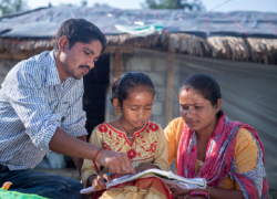 A father and mother read with their daughter in front of their home in Nepal. 