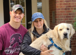 A young couple poses in front of a home. The woman is holding a large vanilla lab.