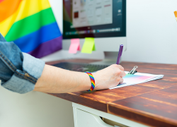 A worker wearing a rainbow bracelet writes notes on a piece of paper at a desk. A Pride flag is displayed next to their computer.