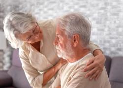 An older woman leans toward an older man who is seated. Her arms enfold him supportively.
