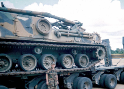 A woman in military fatigues stands in front of an enormous military vehicle.