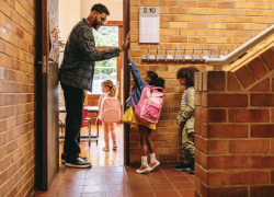 A teacher stands in the doorway of a classroom, giving high fives to young children as they enter.