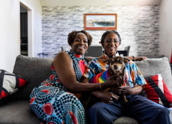 Two individuals smiling and sitting on a sofa in a cozy living room, holding a small dog wearing a rainbow bandana. The room is decorated with a brick-patterned wall and modern furnishings.