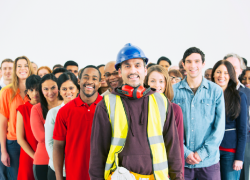 A man wearing a hardhat, ear protection and a high-visibility vest stands in front of a diverse group of smiling workers. 