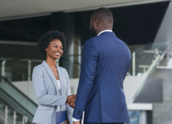 A young woman with a blue blazer smiles while shaking the hand of a man in a blue suit. They are standing in a modern office lobby. 