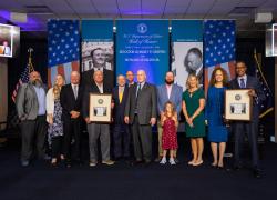 A group of family members and government officials attend a ceremony to induct Sen. Robert Griffin and Howard Jenkins Jr. into the Hall of Honor. Banners behind them have the American flag and black and white photos of both men.