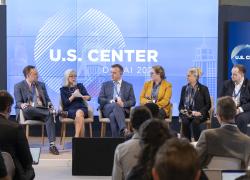 Thea Lee holds a microphone while speaking during a panel discussion with five other people during COP28. The backdrop says “U.S. Center, Dubai 2023.”